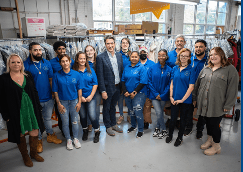 A group of people, some in blue shirts and others in casual attire, stand together in a dry-cleaning facility with racks of clothes in the background.