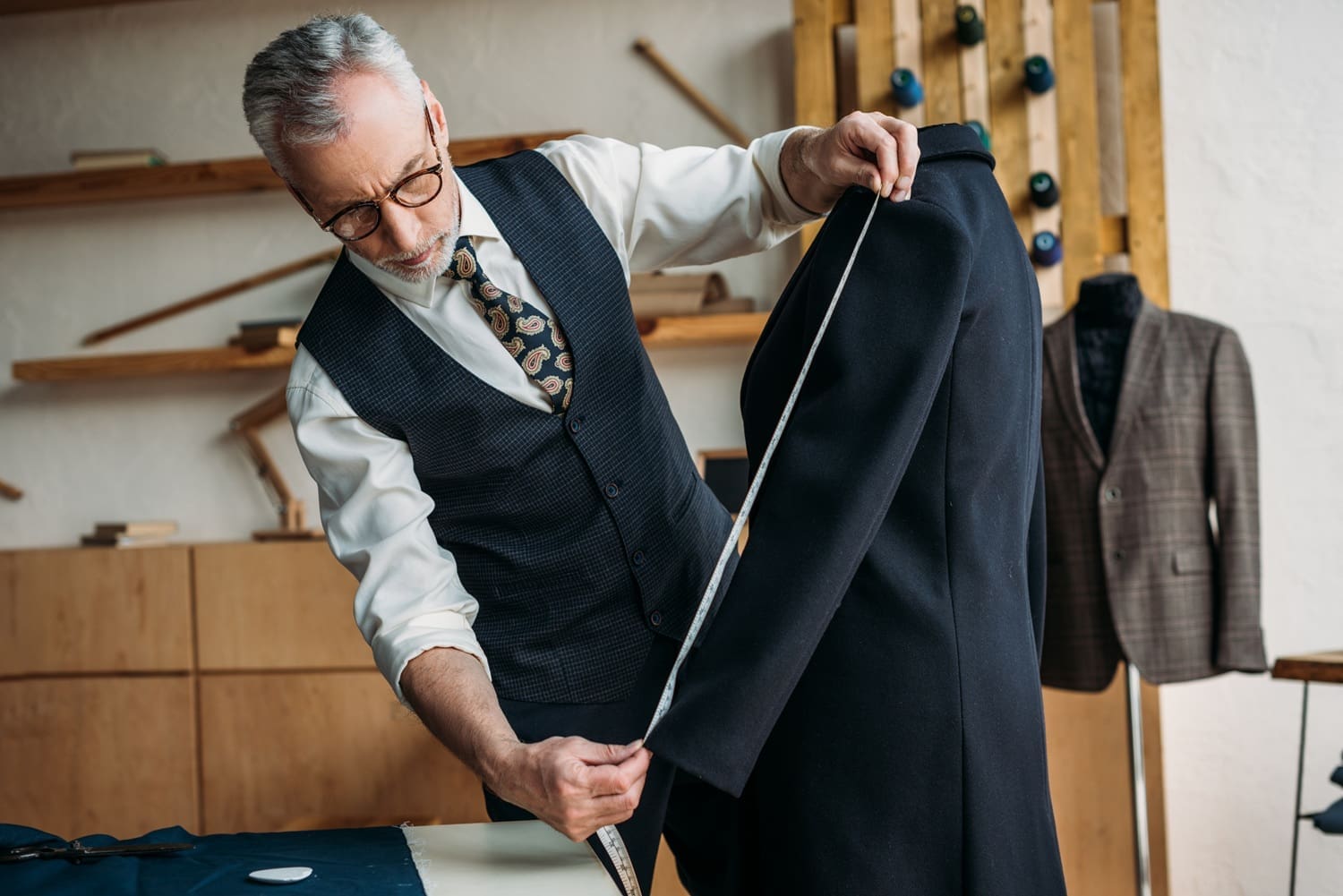 A tailor in a vest measures the sleeve of a black jacket on a dress form in a workshop, with fabric and tools visible in the background.