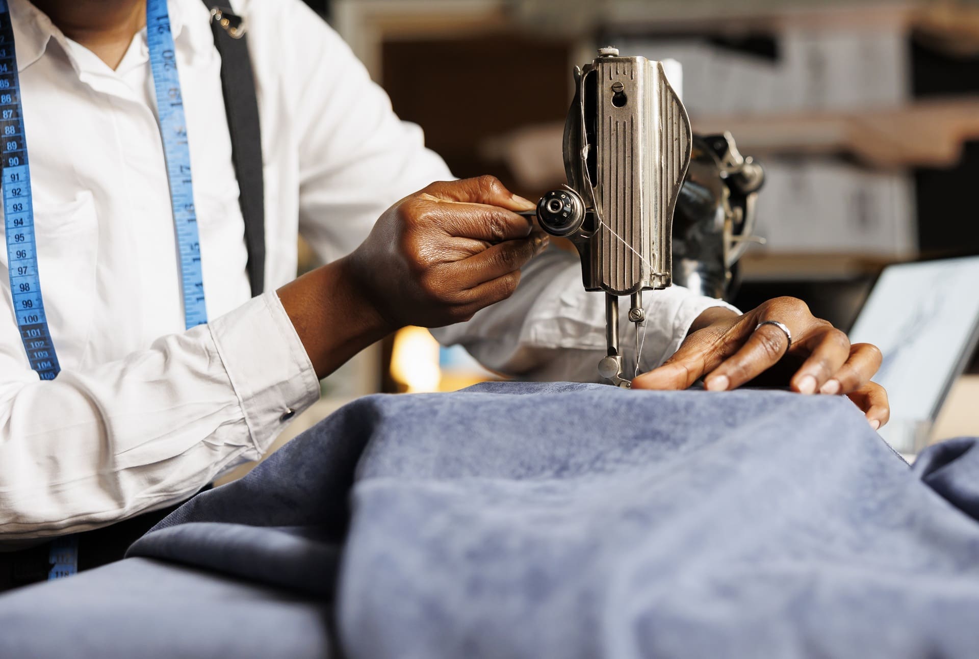 A person operates a sewing machine, stitching blue fabric, with a blue measuring tape draped around their neck.