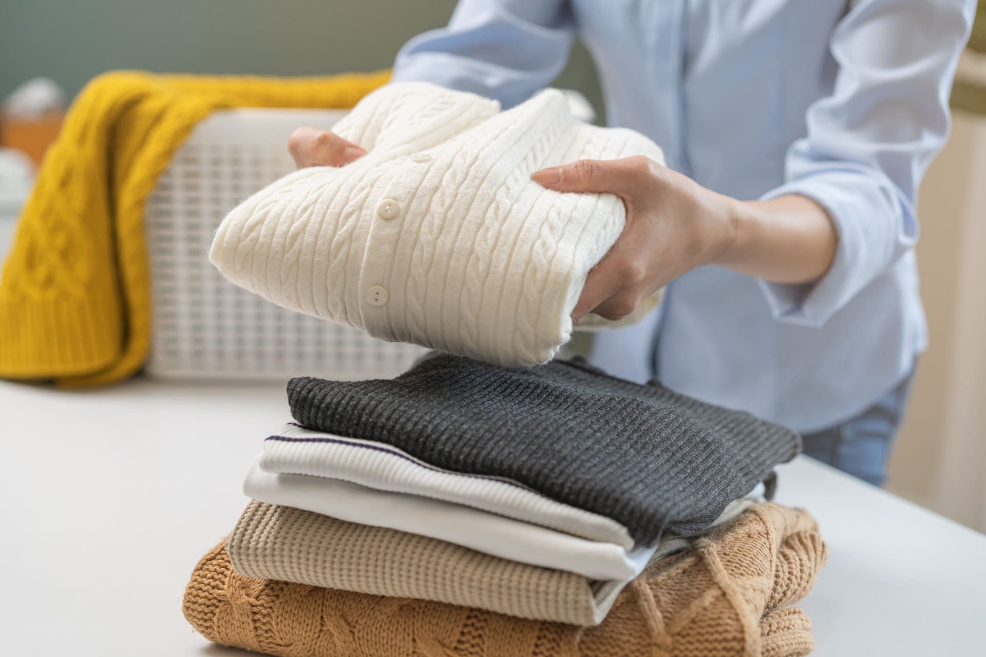 A person folds a white knit sweater and places it on a stack of other folded sweaters on a table.