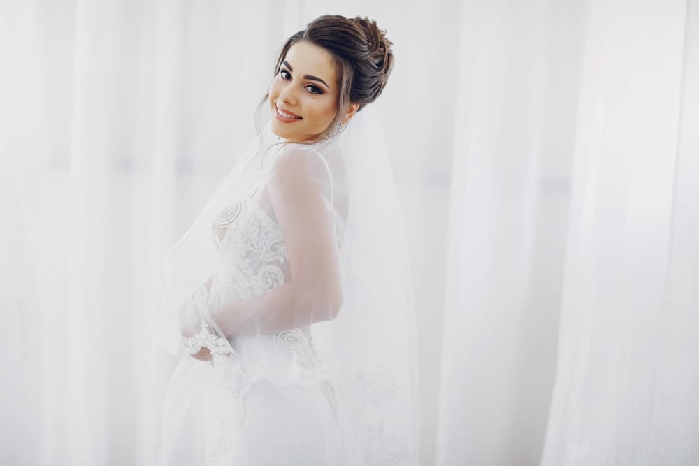 A bride in a white wedding dress and veil stands in front of white curtains, smiling and looking over her shoulder.