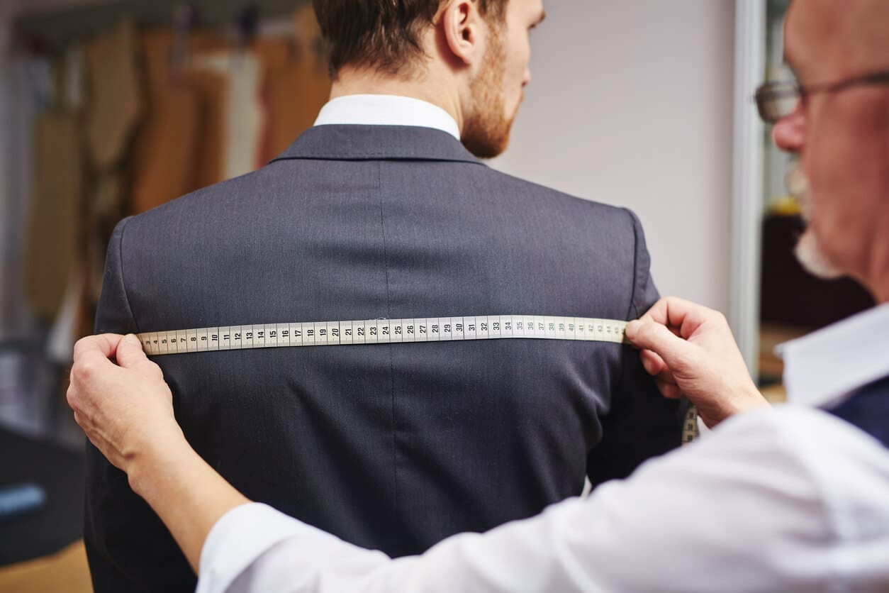 A tailor measures the back width of a man wearing a suit jacket using a measuring tape.